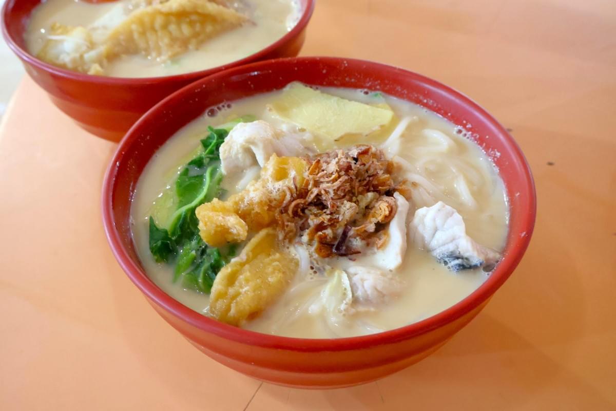A red bowl filled with rich, creamy fish soup noodle dish topped with fresh fish slices, fried fish pieces, green vegetables, noodles, and crispy fried shallots. Another bowl of the milky fish soup sits partially visible in the background on an orange table. A red bowl filled with rich, creamy fish soup noodle dish topped with fresh fish slices, fried fish pieces, green vegetables, noodles, and crispy fried shallots. Another bowl of the milky fish soup sits partially visible in the background on an orange table.
