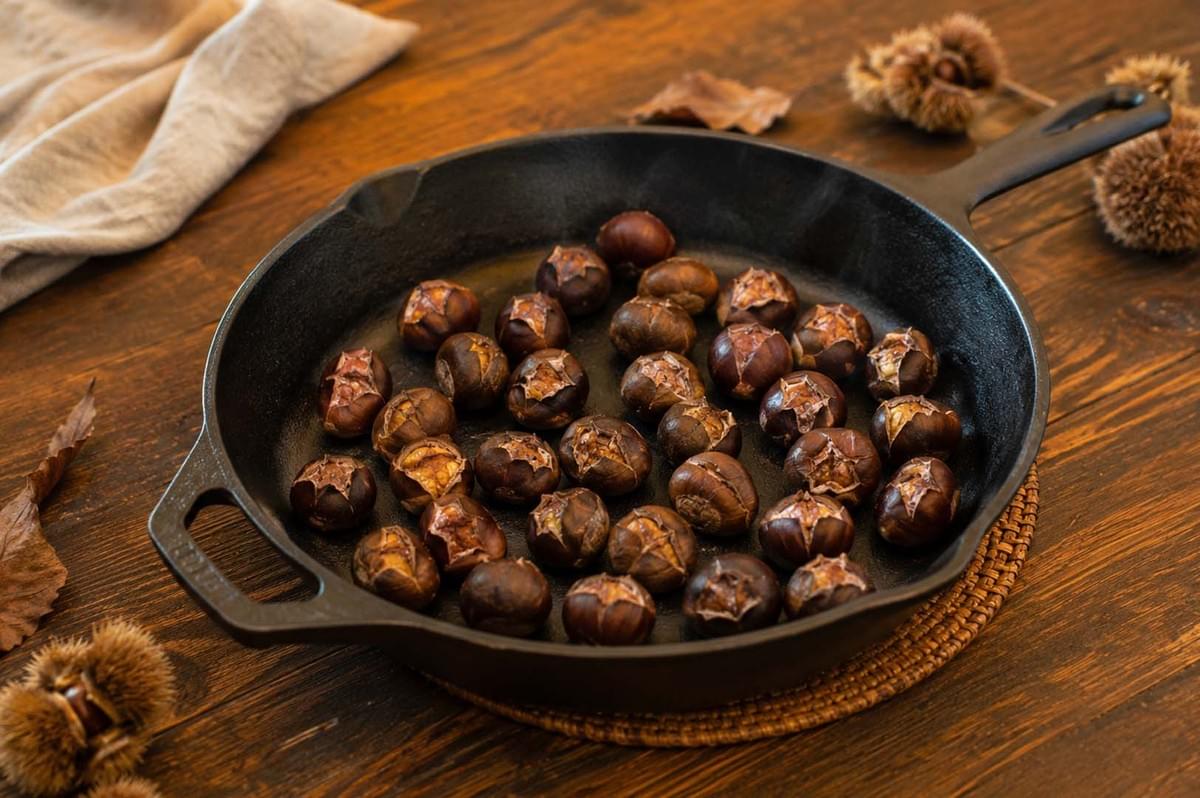 A black cast iron skillet filled with roasted chestnuts, their shells split open to reveal the cooked nut inside A black cast iron skillet filled with roasted chestnuts, their shells split open to reveal the cooked nut inside