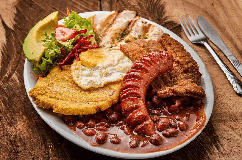 A full plate of Bandeja Paisa featuring red beans in sauce, a large grilled chorizo, crispy chicharrón, a fried egg, a golden patacón (fried plantain), and a side salad with avocado. A full plate of Bandeja Paisa featuring red beans in sauce, a large grilled chorizo, crispy chicharrón, a fried egg, a golden patacón (fried plantain), and a side salad with avocado.