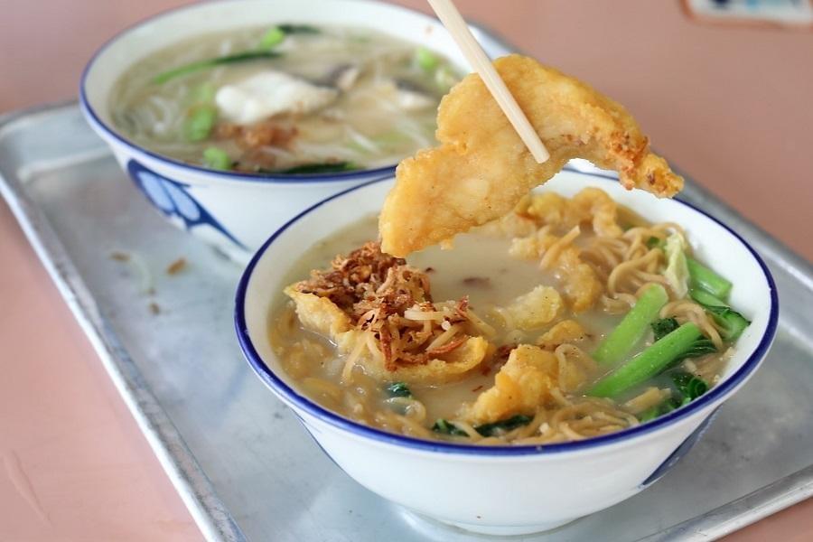 Chopsticks hold a piece of golden fried fish above a bowl of noodle soup with leafy greens and crispy shallots. Another bowl of fish soup rests on the metal tray in the background. Chopsticks hold a piece of golden fried fish above a bowl of noodle soup with leafy greens and crispy shallots. Another bowl of fish soup rests on the metal tray in the background.