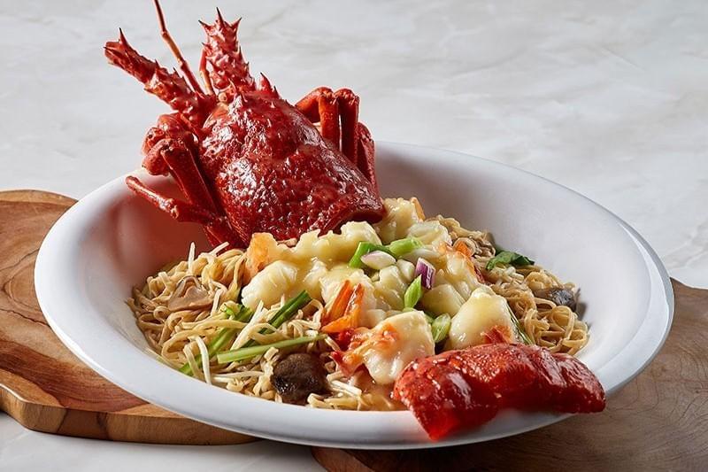 A bright red cooked spiny lobster rests in a large white bowl served atop stir-fried noodles with meat and vegetables. The dish is garnished with sliced green onions and sits on a wooden board against a light marble background. A bright red cooked spiny lobster rests in a large white bowl served atop stir-fried noodles with meat and vegetables. The dish is garnished with sliced green onions and sits on a wooden board against a light marble background.