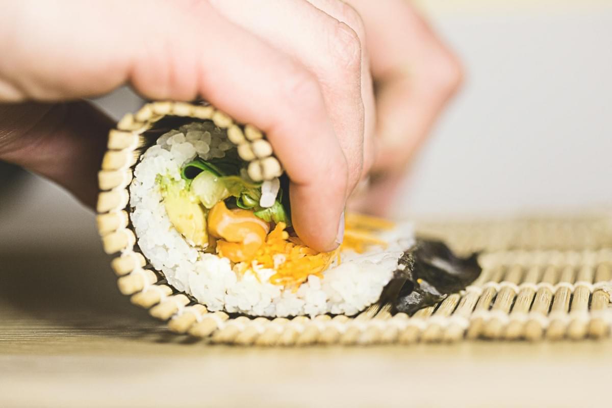 A person's hands use a bamboo rolling mat to shape a homemade sushi roll tightly. The cross-section reveals sushi rice, nori, greens, avocado, shredded carrots, and a creamy sauce. A person's hands use a bamboo rolling mat to shape a homemade sushi roll tightly. The cross-section reveals sushi rice, nori, greens, avocado, shredded carrots, and a creamy sauce.