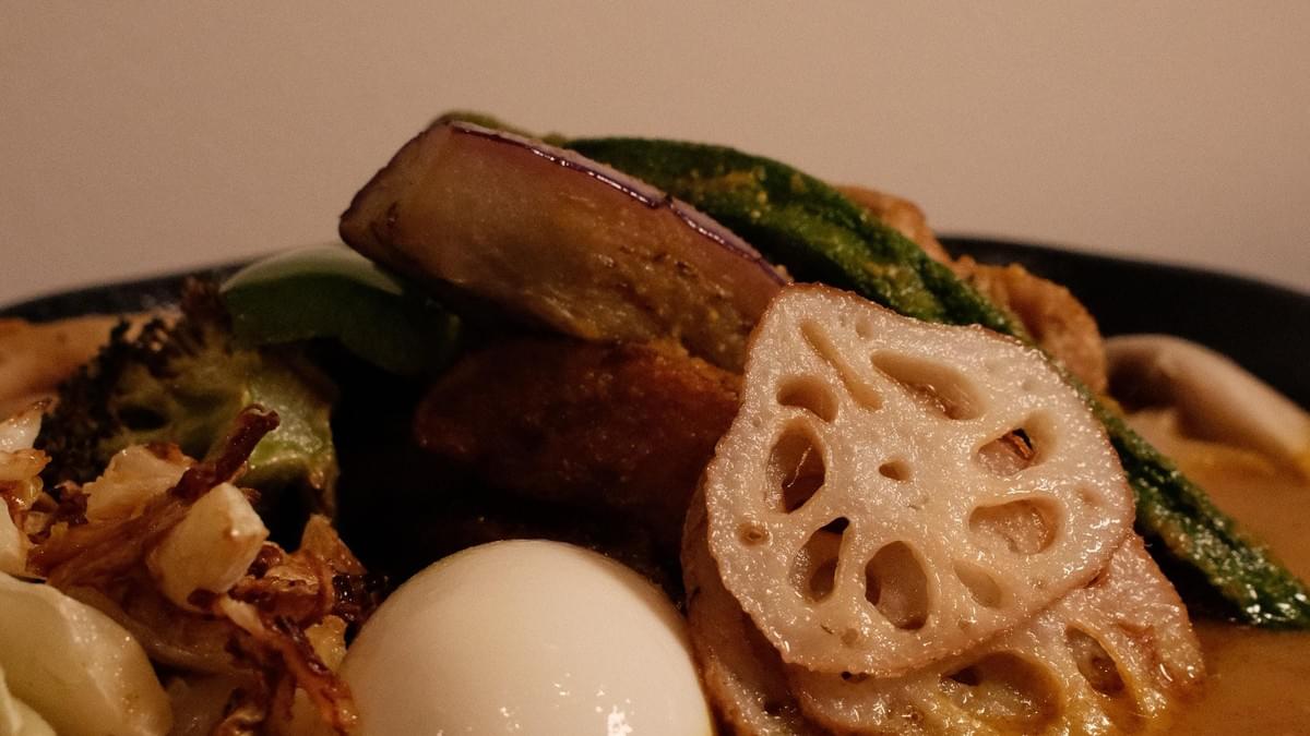 A close-up shot of a savory soup curry dish topped with cooked vegetables and a soft-boiled egg. Prominently featured in the foreground are sliced lotus root, roasted eggplant, okra, and broccoli resting in a warm broth. A close-up shot of a savory soup curry dish topped with cooked vegetables and a soft-boiled egg. Prominently featured in the foreground are sliced lotus root, roasted eggplant, okra, and broccoli resting in a warm broth.