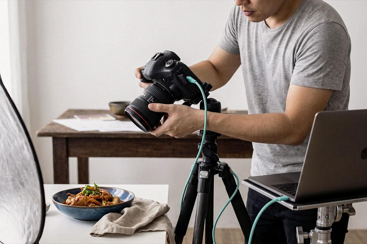 Food photographer adjusting a DSLR camera on a tripod while photographing a plated dish, with studio lighting and a laptop nearby. Food photographer adjusting a DSLR camera on a tripod while photographing a plated dish, with studio lighting and a laptop nearby.