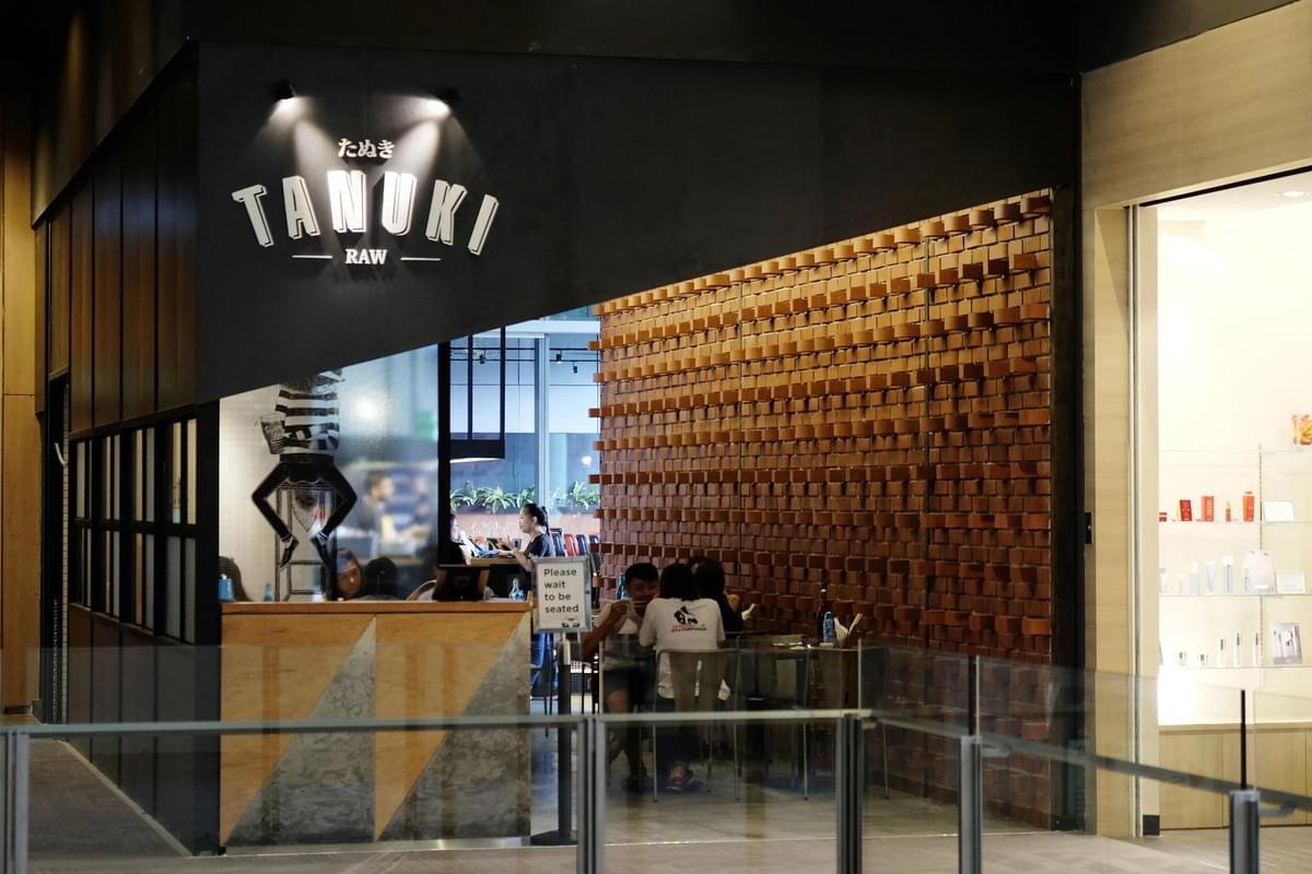 The exterior of a restaurant named "Tanuki Raw" features a modern black facade with white illuminated signage above a reception counter. Inside, patrons sit at tables alongside a textured feature wall made of protruding brown bricks. The exterior of a restaurant named "Tanuki Raw" features a modern black facade with white illuminated signage above a reception counter. Inside, patrons sit at tables alongside a textured feature wall made of protruding brown bricks.