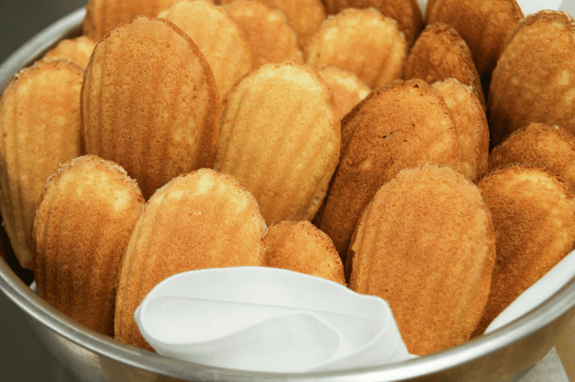 A large metal bowl overflowing with perfectly baked, golden-brown madeleines, showcasing their distinct ridged shell patterns. A large metal bowl overflowing with perfectly baked, golden-brown madeleines, showcasing their distinct ridged shell patterns.