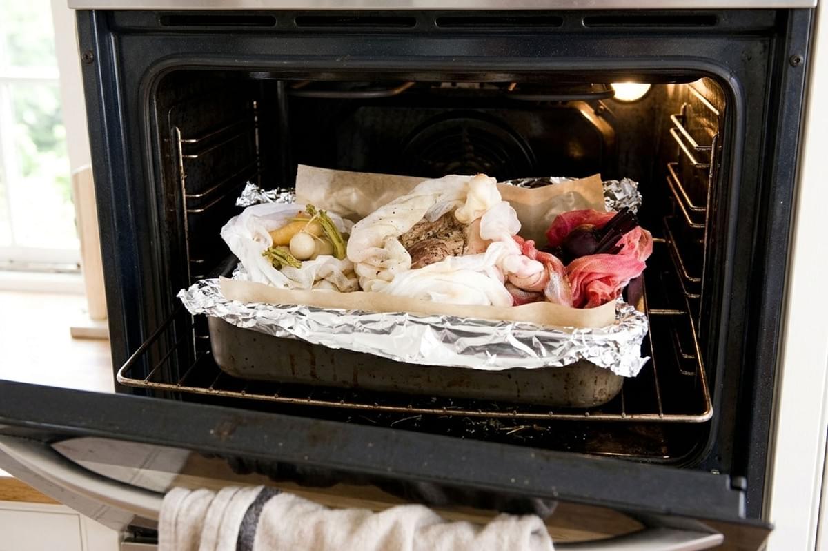 Roasting pan inside an oven containing meat and vegetables wrapped in cloth and foil for slow cooking Roasting pan inside an oven containing meat and vegetables wrapped in cloth and foil for slow cooking