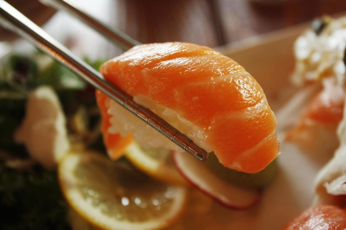 A pair of metal chopsticks holds a fresh piece of salmon nigiri, showcasing the glossy orange fish draped over a small bed of white rice. The background is softly blurred, featuring a garnishment of lemon slices, radish, and fresh greens on a white plate. A pair of metal chopsticks holds a fresh piece of salmon nigiri, showcasing the glossy orange fish draped over a small bed of white rice. The background is softly blurred, featuring a garnishment of lemon slices, radish, and fresh greens on a white plate.