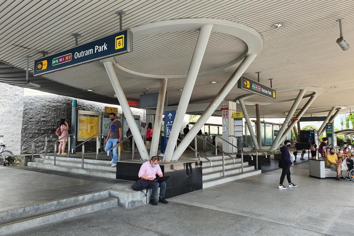 This image shows the modern, sheltered entrance of Outram Park MRT station Exit 6 in Singapore, featuring prominent signage for the East West, North East, and Thomson-East Coast lines. Commuters are seen navigating the area, including a man sitting on the steps using a laptop and others walking toward the station's fare gates. This image shows the modern, sheltered entrance of Outram Park MRT station Exit 6 in Singapore, featuring prominent signage for the East West, North East, and Thomson-East Coast lines. Commuters are seen navigating the area, including a man sitting on the steps using a laptop and others walking toward the station's fare gates.