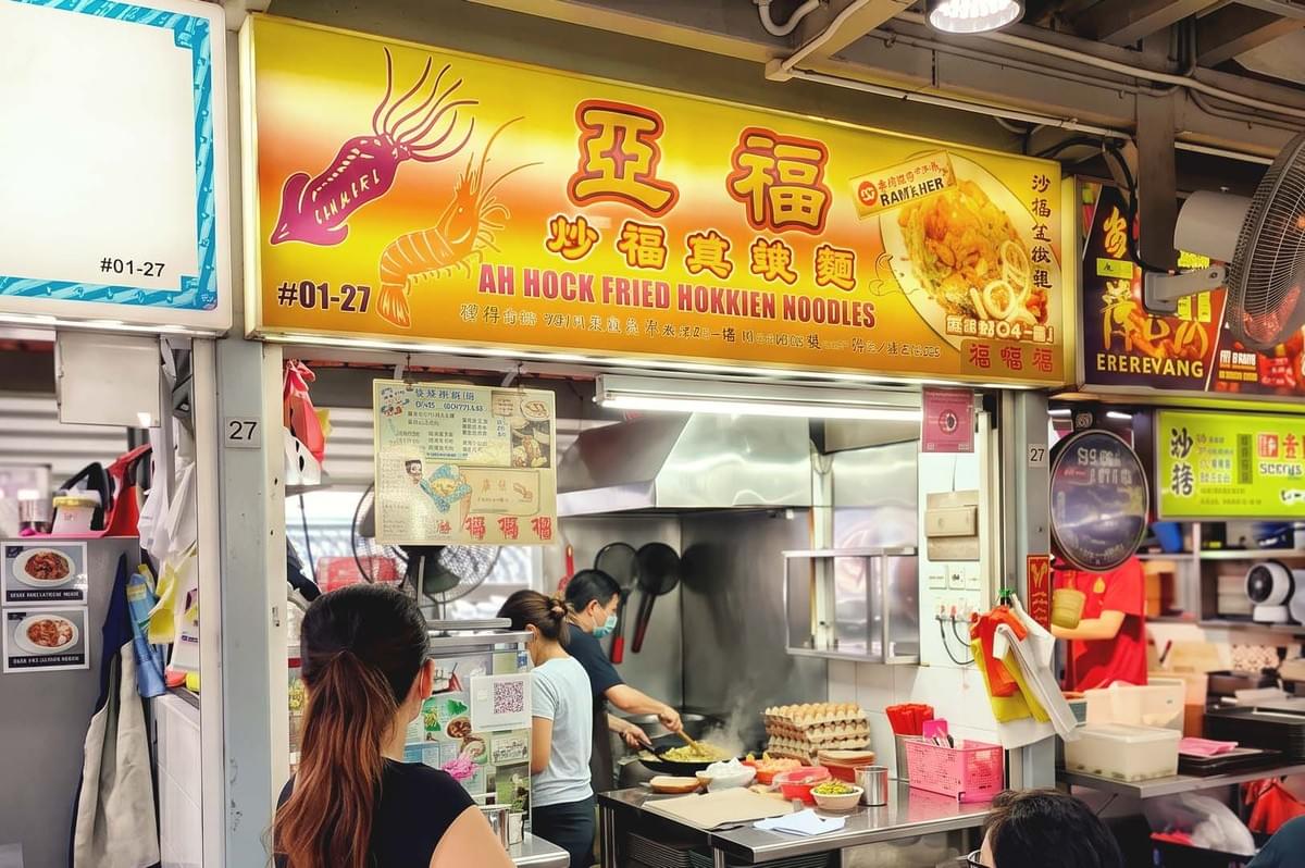 Storefront with yellow orange gradient colored signage of Ah Hok Friend Hokkien Noodles with seafood icons and photo of fried noodles Storefront with yellow orange gradient colored signage of Ah Hok Friend Hokkien Noodles with seafood icons and photo of fried noodles