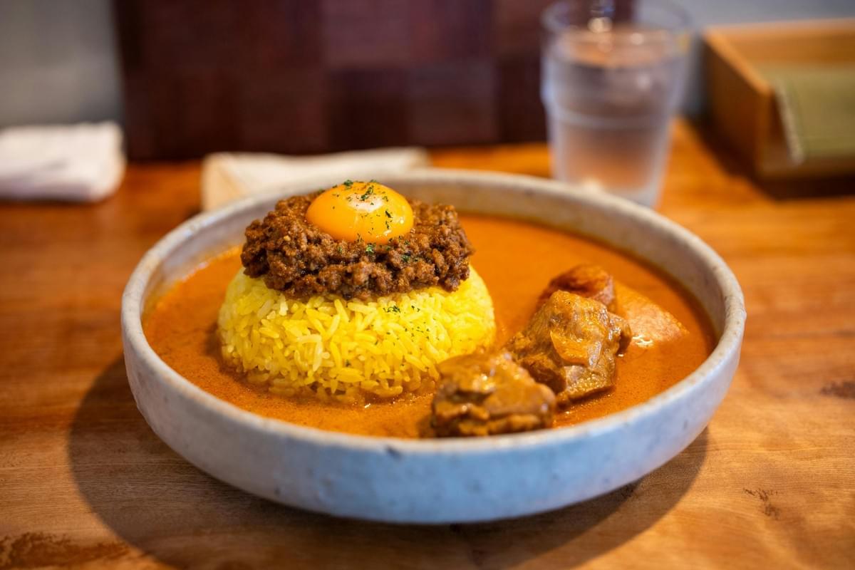 This image features a shallow ceramic bowl containing a mound of yellow rice surrounded by a rich curry sauce and chunks of tender meat. The rice is topped with a layer of savory minced meat and a raw egg yolk, sitting on a wooden table with a glass of water in the background. This image features a shallow ceramic bowl containing a mound of yellow rice surrounded by a rich curry sauce and chunks of tender meat. The rice is topped with a layer of savory minced meat and a raw egg yolk, sitting on a wooden table with a glass of water in the background.