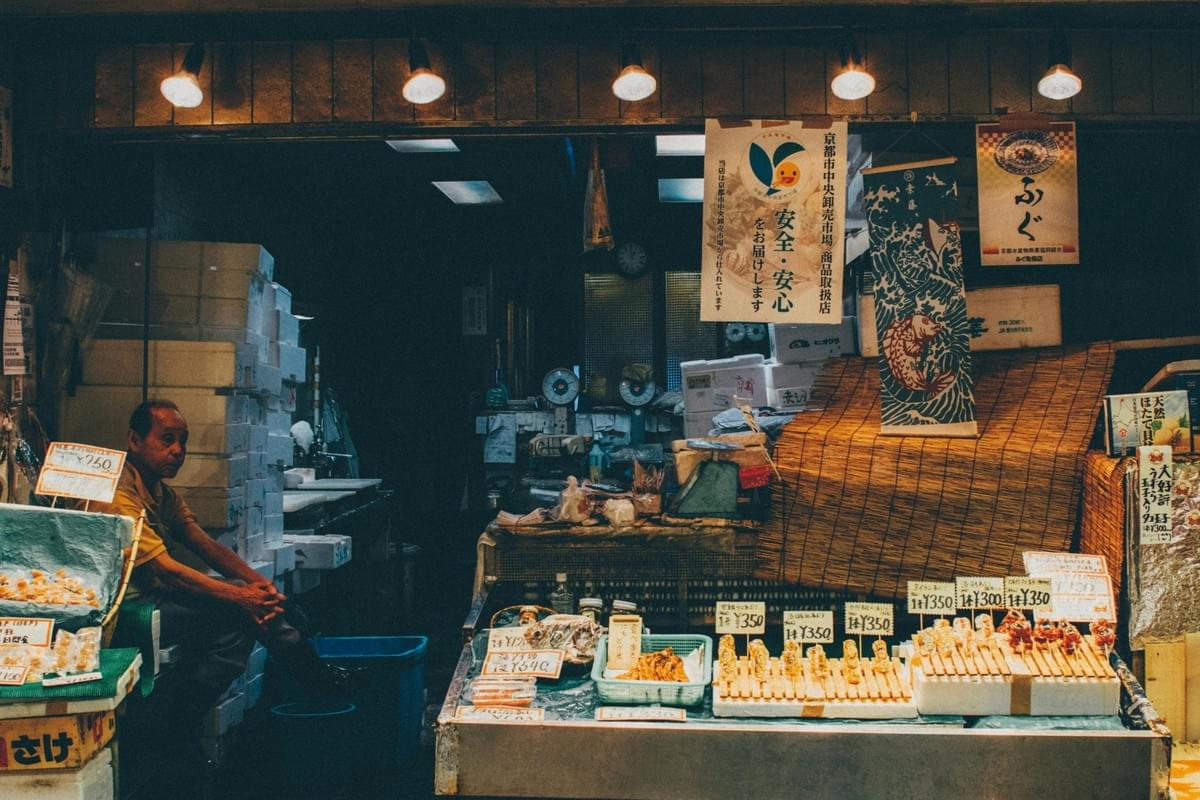 An older man sits quietly beside stacks of white styrofoam boxes at a traditional market stall illuminated by warm overhead lights. The counter in front displays neatly arranged skewers of food and packaged items, marked with handwritten price tags in Japanese currency. An older man sits quietly beside stacks of white styrofoam boxes at a traditional market stall illuminated by warm overhead lights. The counter in front displays neatly arranged skewers of food and packaged items, marked with handwritten price tags in Japanese currency.
