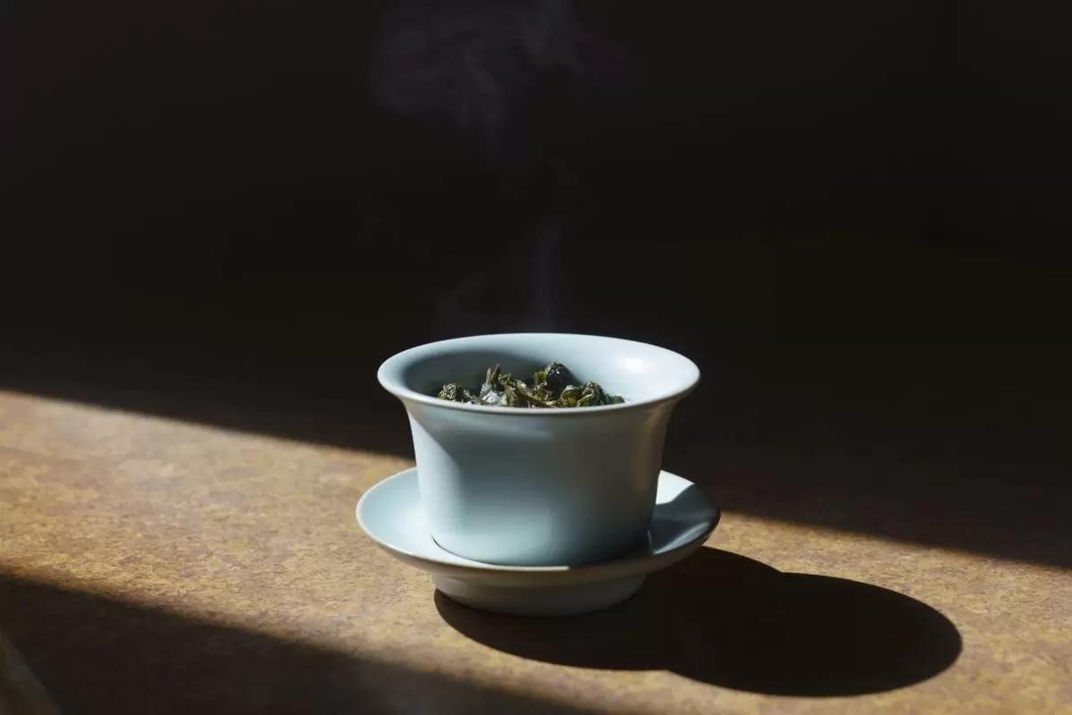 A light blue ceramic gaiwan filled with steeped green tea leaves rests on a matching saucer, illuminated by a beam of sunlight. Delicate wisps of steam rise from the hot tea against a dramatic, dark background. A light blue ceramic gaiwan filled with steeped green tea leaves rests on a matching saucer, illuminated by a beam of sunlight. Delicate wisps of steam rise from the hot tea against a dramatic, dark background.