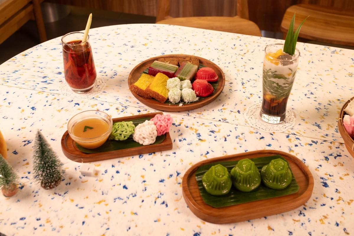 A table displays an assortment of traditional Indonesian or Southeast Asian desserts and drinks served on wooden platters and in glass dishes. The spread includes colorful kue, klepon, a bowl of sauce with noodles, and iced beverages garnished with lemongrass and pandan leaves. A table displays an assortment of traditional Indonesian or Southeast Asian desserts and drinks served on wooden platters and in glass dishes. The spread includes colorful kue, klepon, a bowl of sauce with noodles, and iced beverages garnished with lemongrass and pandan leaves.