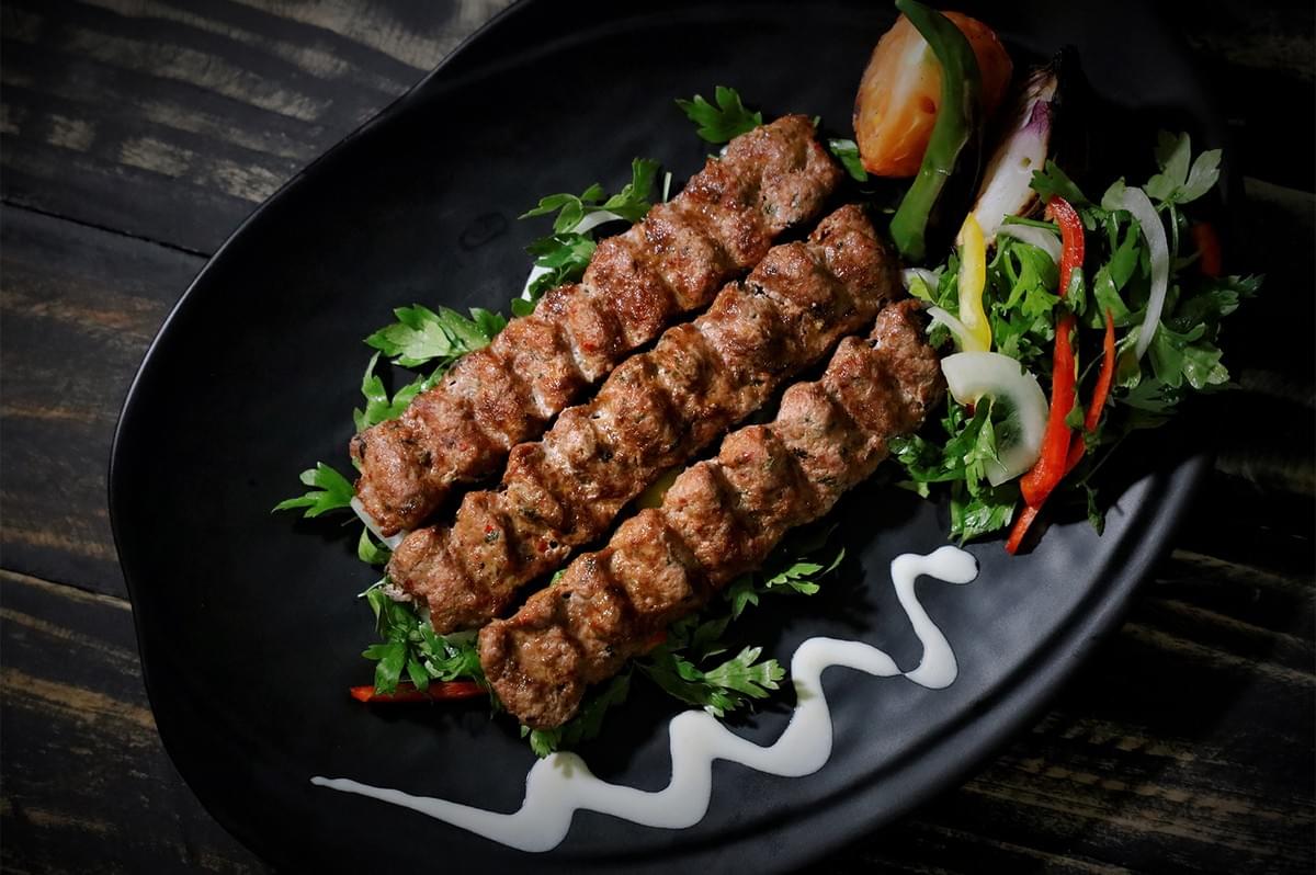 Grilled Adana kebab skewers served with fresh vegetables and yogurt sauce on a black platter. Grilled Adana kebab skewers served with fresh vegetables and yogurt sauce on a black platter.