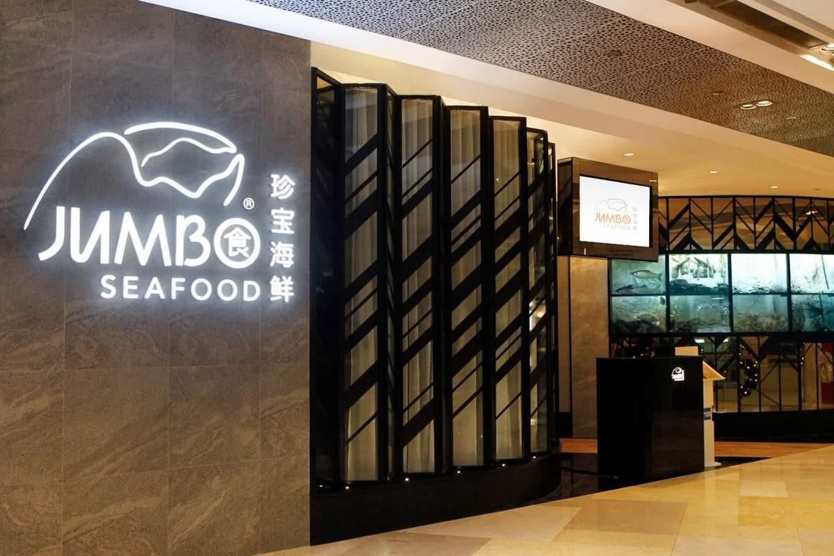 An illuminated sign on a textured wall displays the logo and name "JUMBO SEAFOOD" alongside Chinese characters. A decorative dark panel divider leads toward a reception podium and large fish tanks inside the restaurant entrance. An illuminated sign on a textured wall displays the logo and name "JUMBO SEAFOOD" alongside Chinese characters. A decorative dark panel divider leads toward a reception podium and large fish tanks inside the restaurant entrance.