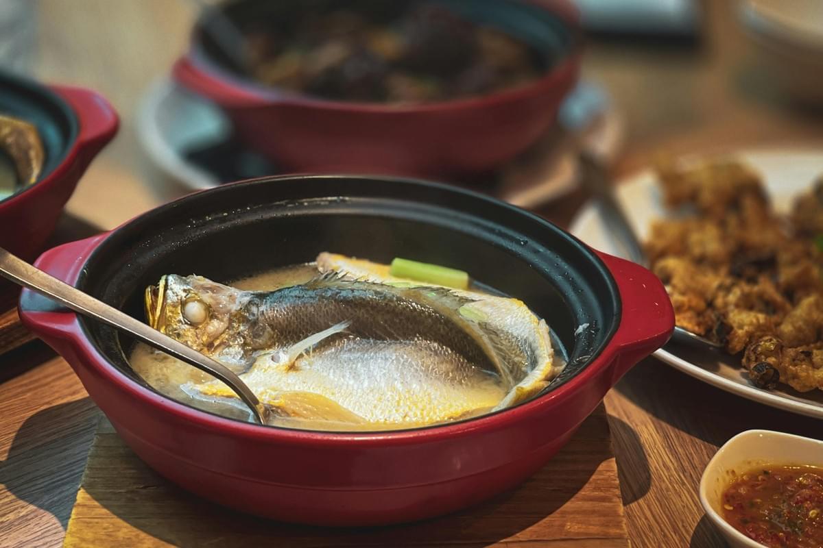 A red clay pot filled with broth and a whole cooked fish rests on a wooden trivet. Other dishes and sauces are scattered around the dining table in soft focus. A red clay pot filled with broth and a whole cooked fish rests on a wooden trivet. Other dishes and sauces are scattered around the dining table in soft focus.