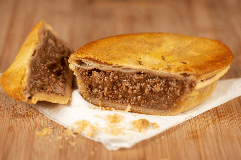 A classic individual Australian meat pie sliced open on a wooden board, showing a moist, dark gravy and minced beef interior with flaky pastry crumbs. A classic individual Australian meat pie sliced open on a wooden board, showing a moist, dark gravy and minced beef interior with flaky pastry crumbs.