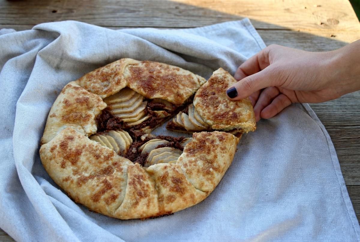 A hand lifting a slice from a baked pear galette resting on a grey cloth napkin over a wooden surface. A hand lifting a slice from a baked pear galette resting on a grey cloth napkin over a wooden surface.
