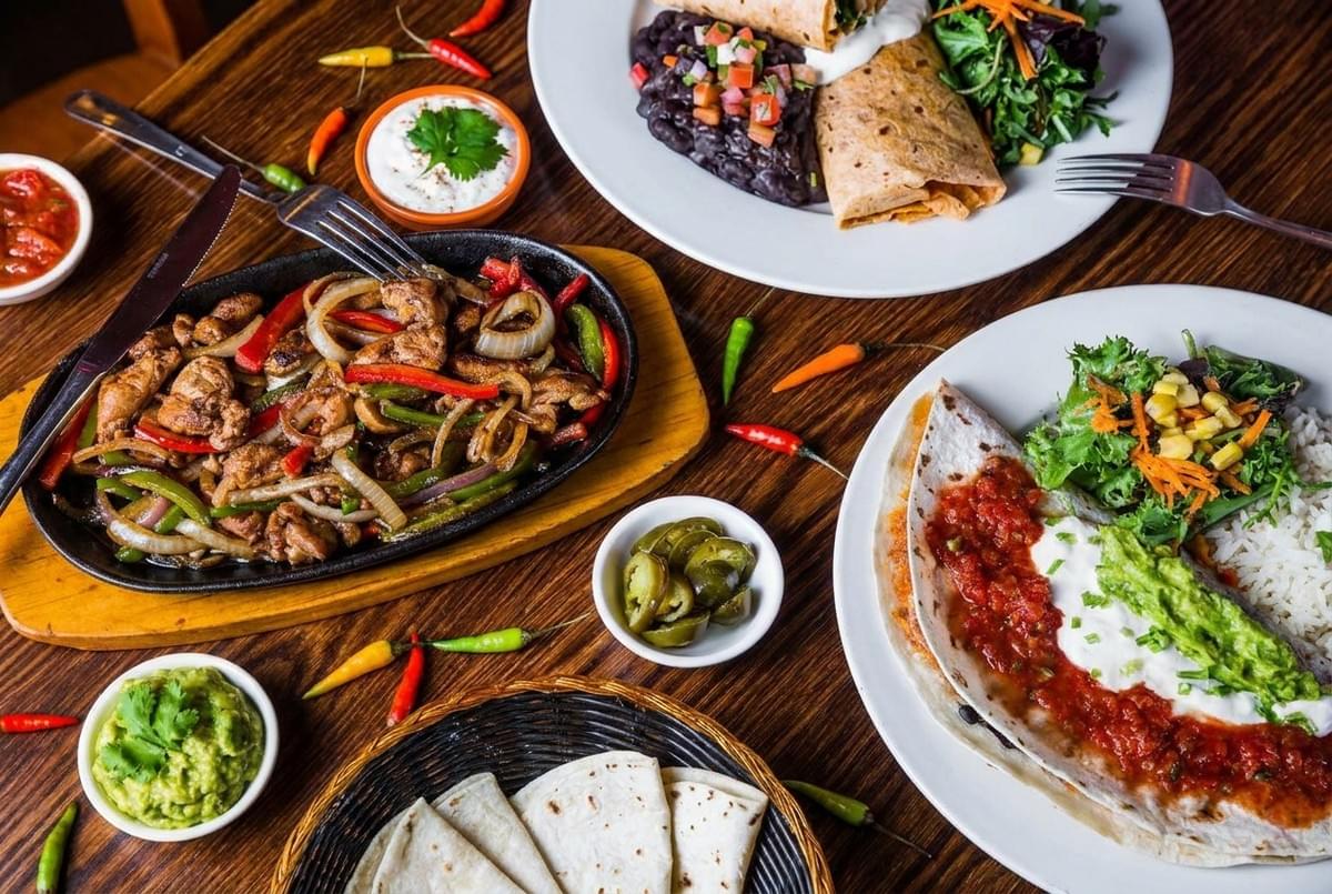 Top-down spread of a Mexican feast featuring sizzling fajitas, burritos, tortillas, salsa, and guacamole on a wooden table. Top-down spread of a Mexican feast featuring sizzling fajitas, burritos, tortillas, salsa, and guacamole on a wooden table.