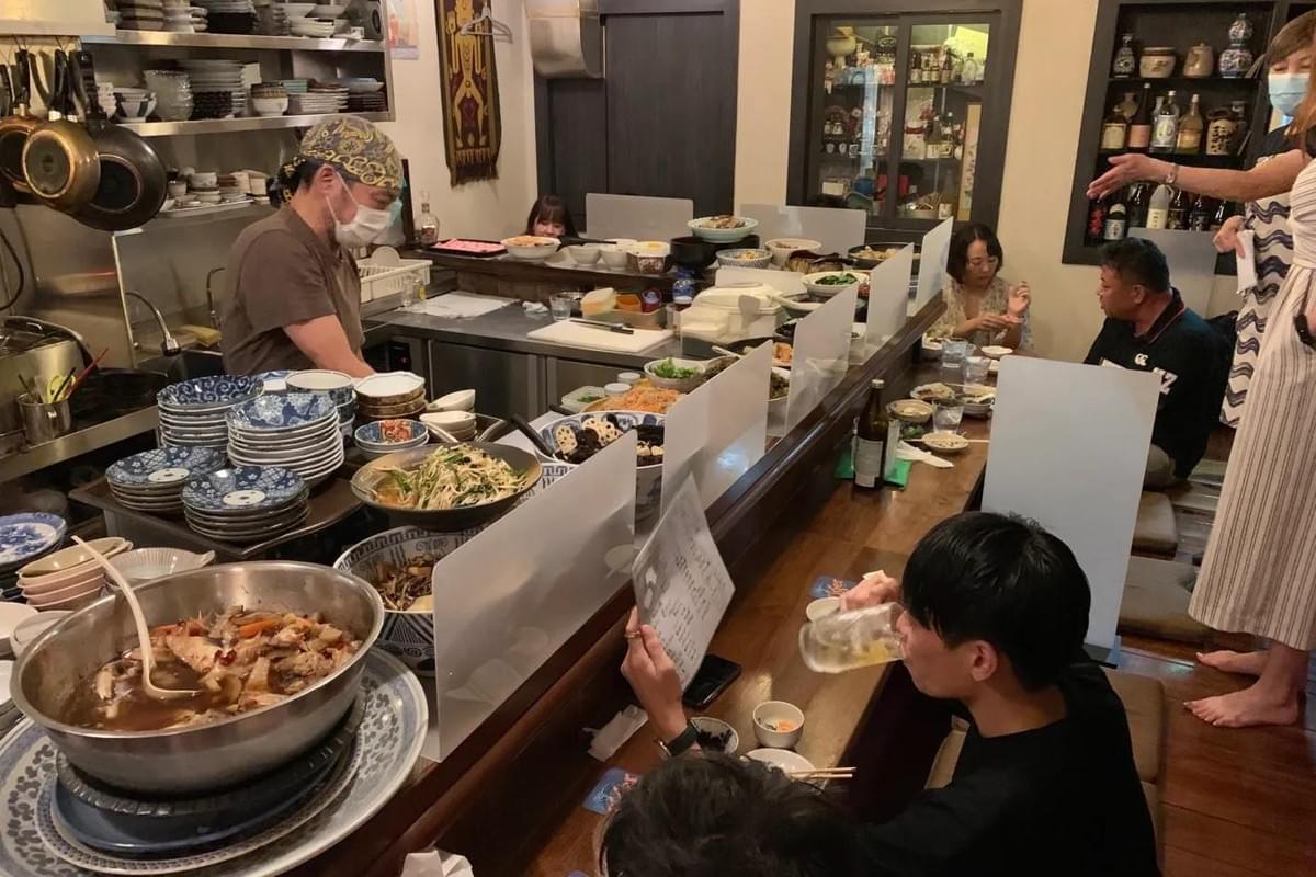 An open kitchen counter at a busy Japanese restaurant is lined with frosted acrylic privacy dividers separating the staff from customers. In the foreground, a customer looks over a menu while taking a drink, as a chef in a face mask prepares food behind stacked plates and large bowls of simmering dishes. An open kitchen counter at a busy Japanese restaurant is lined with frosted acrylic privacy dividers separating the staff from customers. In the foreground, a customer looks over a menu while taking a drink, as a chef in a face mask prepares food behind stacked plates and large bowls of simmering dishes.