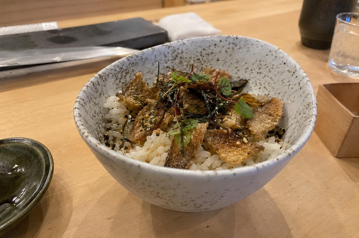 A bowl of white rice is topped with golden-brown, crispy shards of salmon skin, garnished with dark strips of nori, sesame seeds, and small, bright green microgreens. The dish is served in a speckled ceramic bowl, which sits on a smooth wooden tabletop alongside a small dark dish on the left, a glass of water in the background, and a wooden box, suggesting a clean, minimalist dining setting. A bowl of white rice is topped with golden-brown, crispy shards of salmon skin, garnished with dark strips of nori, sesame seeds, and small, bright green microgreens. The dish is served in a speckled ceramic bowl, which sits on a smooth wooden tabletop alongside a small dark dish on the left, a glass of water in the background, and a wooden box, suggesting a clean, minimalist dining setting.