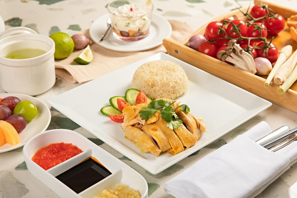 A plate of Hainanese chicken rice sits at the center of a dining table, featuring sliced poached chicken garnished with cilantro, a dome of seasoned rice, and fresh cucumber and tomato slices. The meal is surrounded by various side dishes, including a three-compartment dipping sauce tray, fresh fruit, a small bowl of soup, and a wooden basket filled with cherry tomatoes and mushrooms A plate of Hainanese chicken rice sits at the center of a dining table, featuring sliced poached chicken garnished with cilantro, a dome of seasoned rice, and fresh cucumber and tomato slices. The meal is surrounded by various side dishes, including a three-compartment dipping sauce tray, fresh fruit, a small bowl of soup, and a wooden basket filled with cherry tomatoes and mushrooms