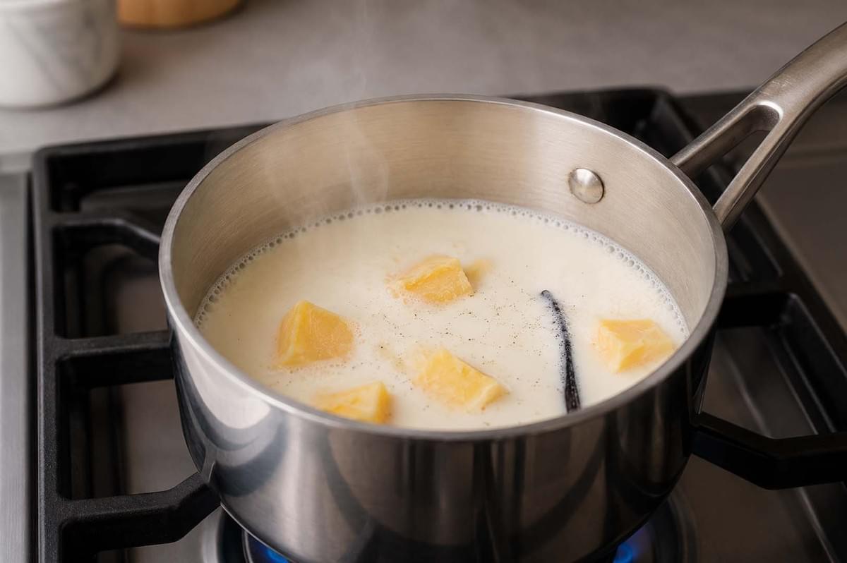 Stainless steel saucepan on a stove heating milk, chunks of butter, and a vanilla bean Stainless steel saucepan on a stove heating milk, chunks of butter, and a vanilla bean