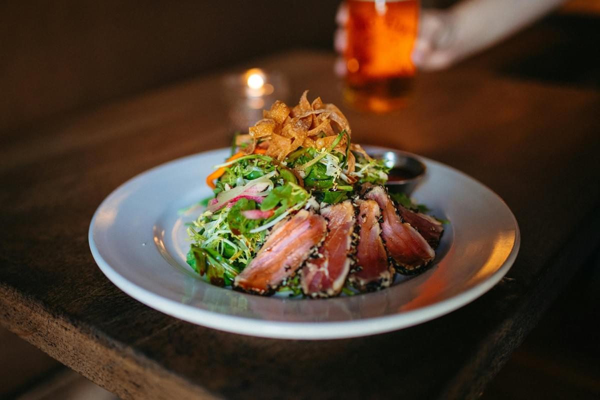 A white plate served on a dark wooden table holds sliced sesame-seared tuna alongside a mixed green salad topped with crispy wonton strips. A glowing candle and a glass of amber beer rest in the softly blurred background. A white plate served on a dark wooden table holds sliced sesame-seared tuna alongside a mixed green salad topped with crispy wonton strips. A glowing candle and a glass of amber beer rest in the softly blurred background.