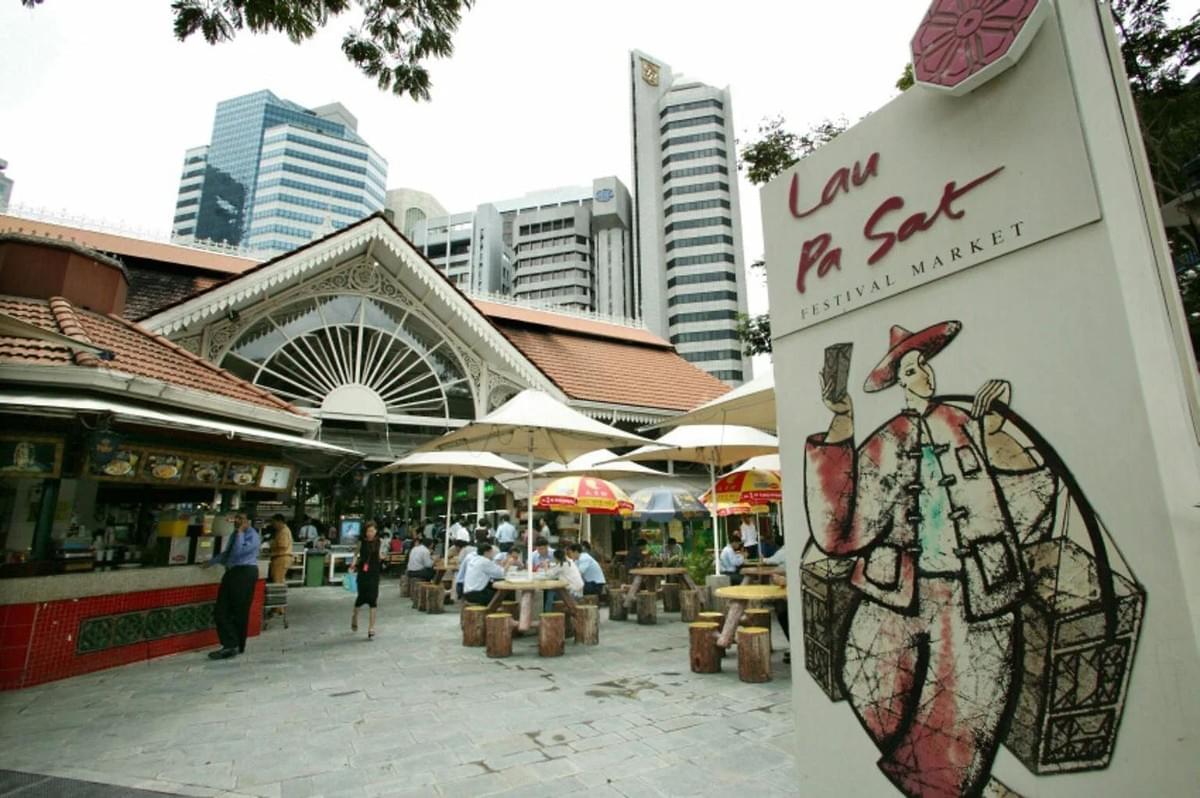 A white sign featuring the text "Lau Pa Sat Festival Market" and an illustration of a traditional vendor stands in the foreground. Behind it, patrons sit at outdoor tables beneath umbrellas outside the historic market hall, framed by modern skyscrapers in the background. A white sign featuring the text "Lau Pa Sat Festival Market" and an illustration of a traditional vendor stands in the foreground. Behind it, patrons sit at outdoor tables beneath umbrellas outside the historic market hall, framed by modern skyscrapers in the background.