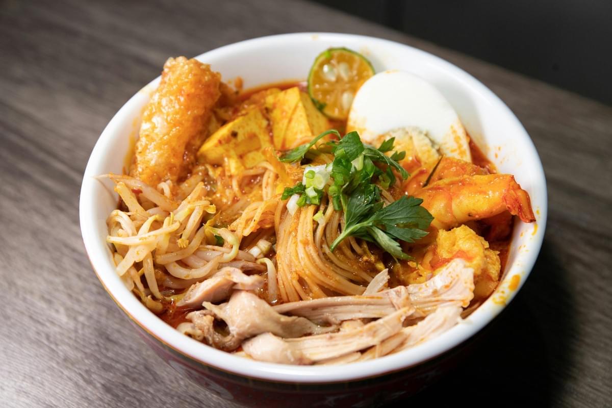 In the image, a white bowl filled with Laksa noodles sits on a dark wooden table. The dish features rice noodles in a rich, reddish curry broth topped with shredded chicken, shrimp, bean sprouts, hard-boiled egg, tofu puff, and fresh herbs. In the image, a white bowl filled with Laksa noodles sits on a dark wooden table. The dish features rice noodles in a rich, reddish curry broth topped with shredded chicken, shrimp, bean sprouts, hard-boiled egg, tofu puff, and fresh herbs.