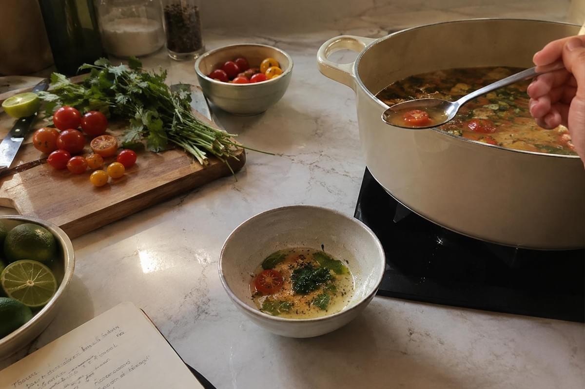 A hand holds a metal spoon tasting soup from a large, off-white enameled Dutch oven set on a stovetop. Nearby on the marble countertop sits a small tasting bowl with broth, herbs, and sliced cherry tomatoes, an open handwritten recipe notebook, and a wooden cutting board scattered with fresh cilantro stems, whole and halved cherry tomatoes, and sliced limes. A hand holds a metal spoon tasting soup from a large, off-white enameled Dutch oven set on a stovetop. Nearby on the marble countertop sits a small tasting bowl with broth, herbs, and sliced cherry tomatoes, an open handwritten recipe notebook, and a wooden cutting board scattered with fresh cilantro stems, whole and halved cherry tomatoes, and sliced limes.