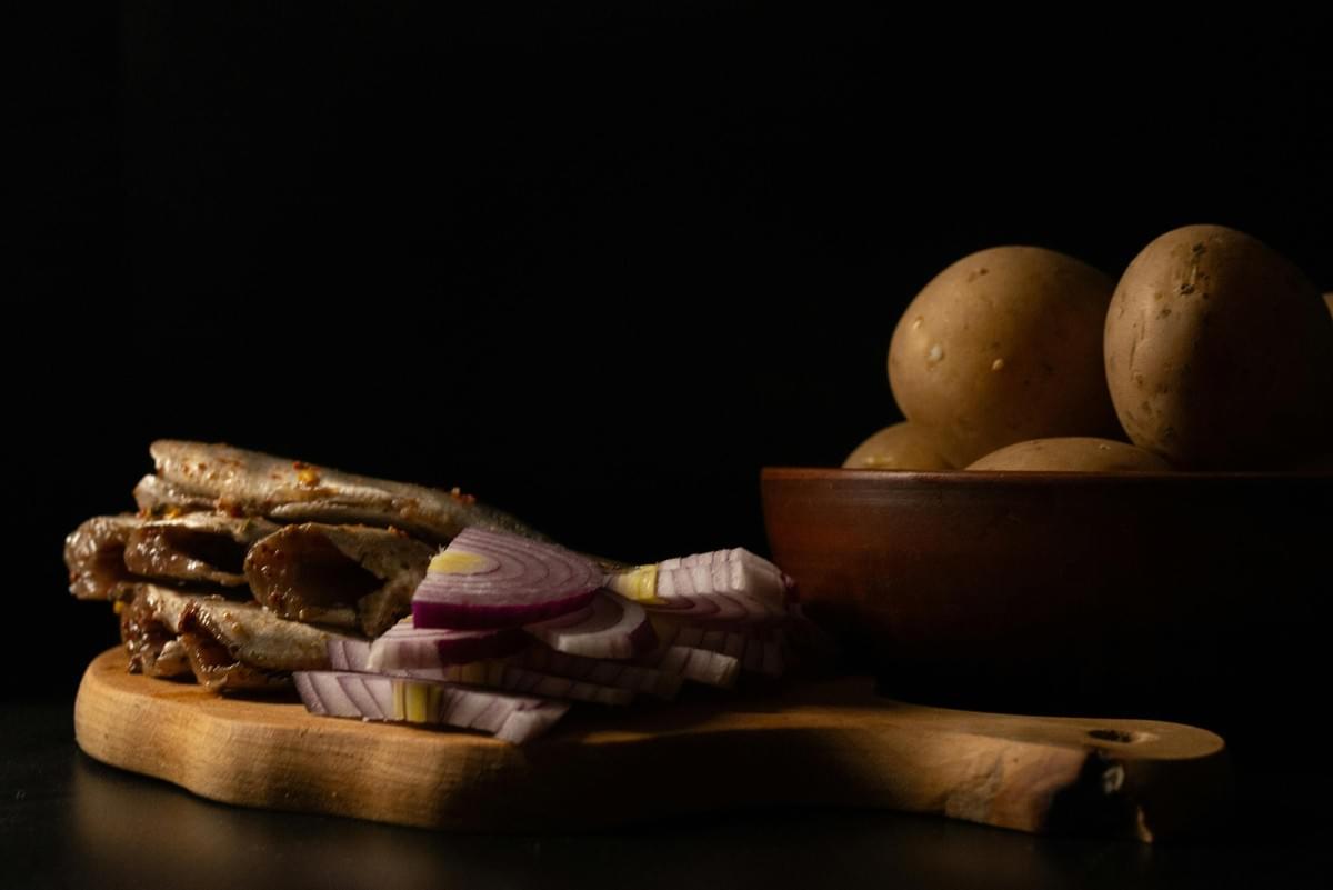 A wooden cutting board holds a stack of seasoned small fish alongside several slices of raw red onion. Next to the board, a brown ceramic bowl is filled with whole, unpeeled potatoes against a solid black background. A wooden cutting board holds a stack of seasoned small fish alongside several slices of raw red onion. Next to the board, a brown ceramic bowl is filled with whole, unpeeled potatoes against a solid black background.