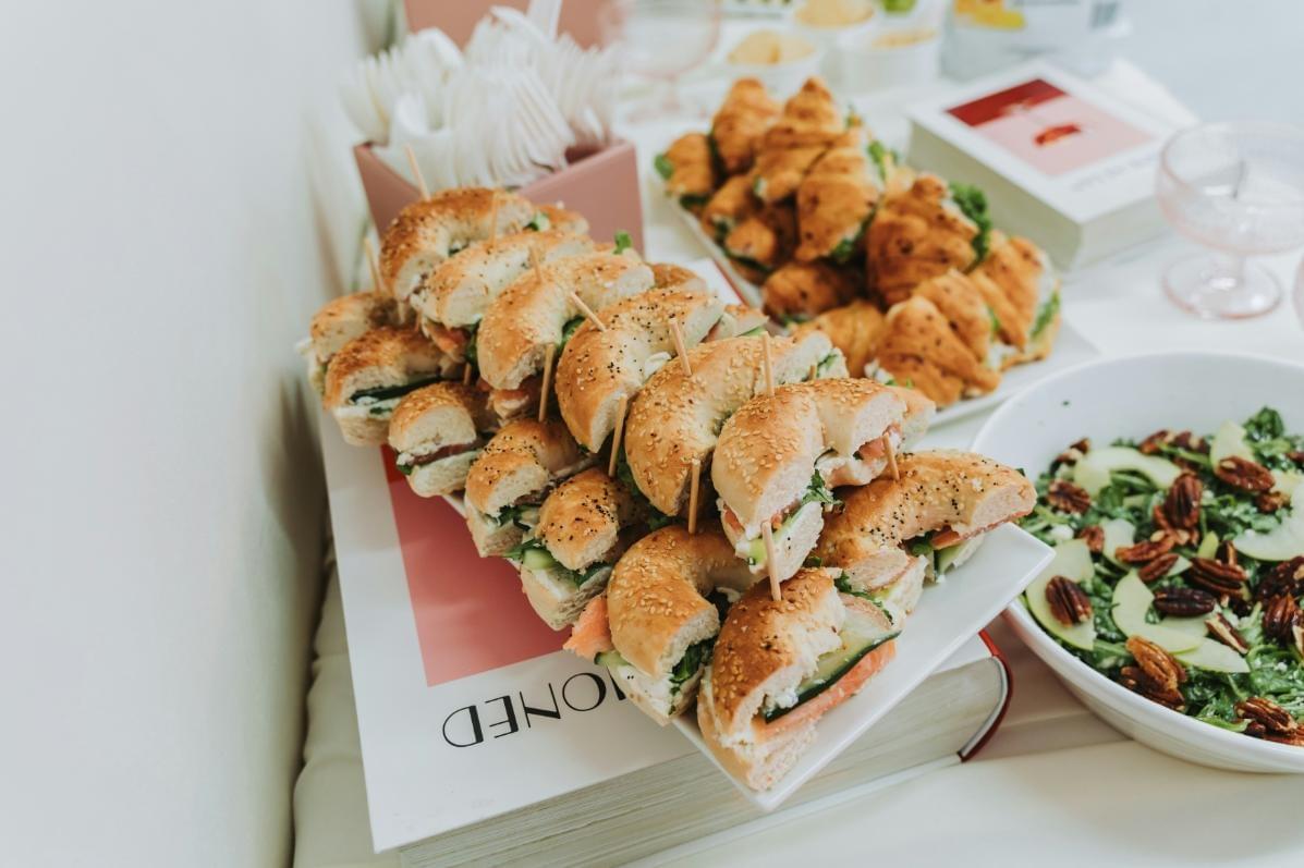 In the image, a catering spread features a platter of assorted bagel sandwich halves held together with toothpicks alongside a tray of mini croissants. Next to the sandwiches, a large white bowl holds a fresh green salad topped with sliced apples and pecans. In the image, a catering spread features a platter of assorted bagel sandwich halves held together with toothpicks alongside a tray of mini croissants. Next to the sandwiches, a large white bowl holds a fresh green salad topped with sliced apples and pecans.