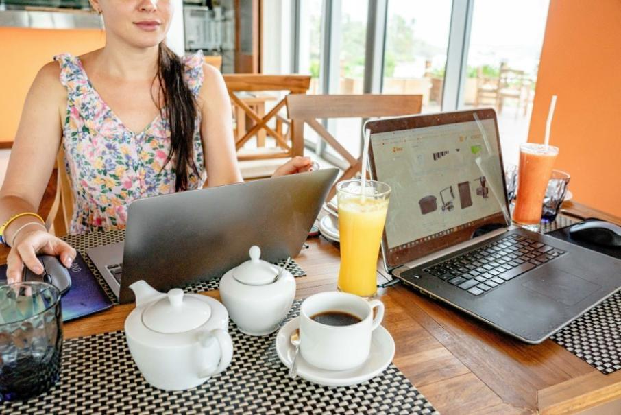 In the image, a woman in a floral top sits at a wooden table working on a laptop with a computer mouse. The table is also set with a second laptop, a cup of coffee, a teapot, a sugar bowl, and glasses of smoothies. In the image, a woman in a floral top sits at a wooden table working on a laptop with a computer mouse. The table is also set with a second laptop, a cup of coffee, a teapot, a sugar bowl, and glasses of smoothies.