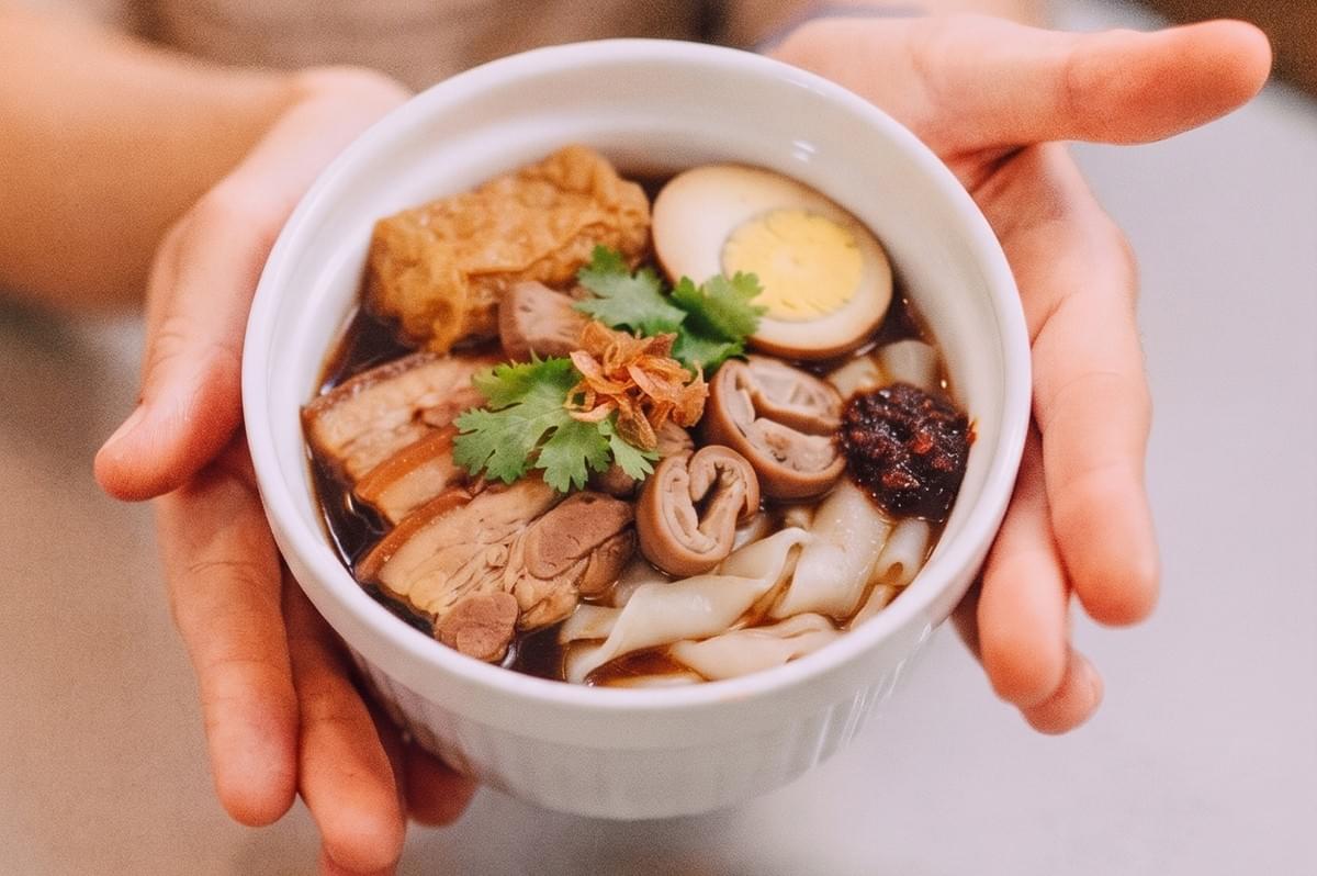 A person holding a bowl of traditional Singaporean kway chap with braised pork, egg, and noodles. A person holding a bowl of traditional Singaporean kway chap with braised pork, egg, and noodles.
