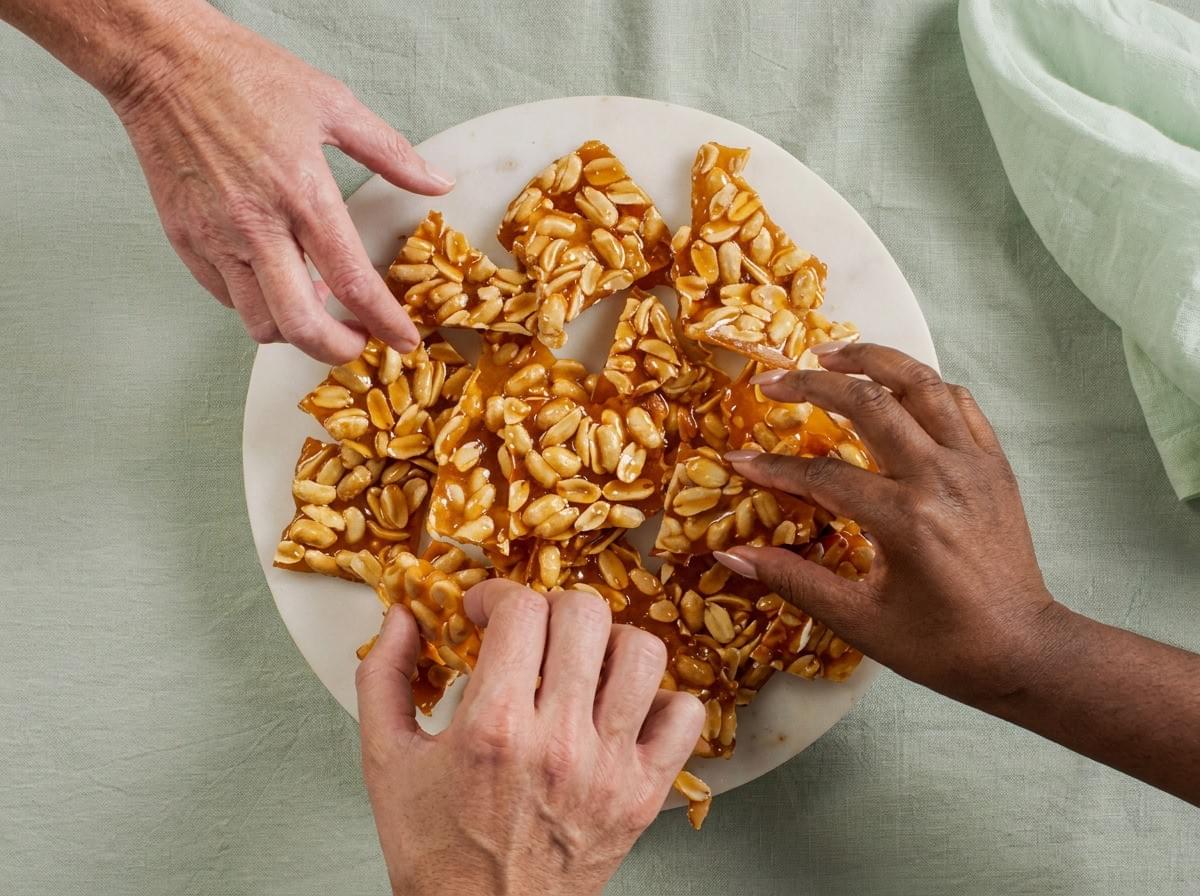 Top-down view of three hands reaching to grab pieces of peanut brittle from a round marble platter on a light green tablecloth. Top-down view of three hands reaching to grab pieces of peanut brittle from a round marble platter on a light green tablecloth.