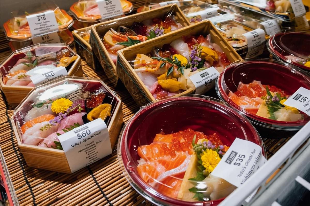 An array of pre-packaged Japanese seafood boxes and bowls on display at a market, featuring price-tagged take-out containers filled with options like salmon and ikura don, signature sushi sets, and deluxe chirashi boxes garnished with yellow blossoms and purple edible flowers. An array of pre-packaged Japanese seafood boxes and bowls on display at a market, featuring price-tagged take-out containers filled with options like salmon and ikura don, signature sushi sets, and deluxe chirashi boxes garnished with yellow blossoms and purple edible flowers.
