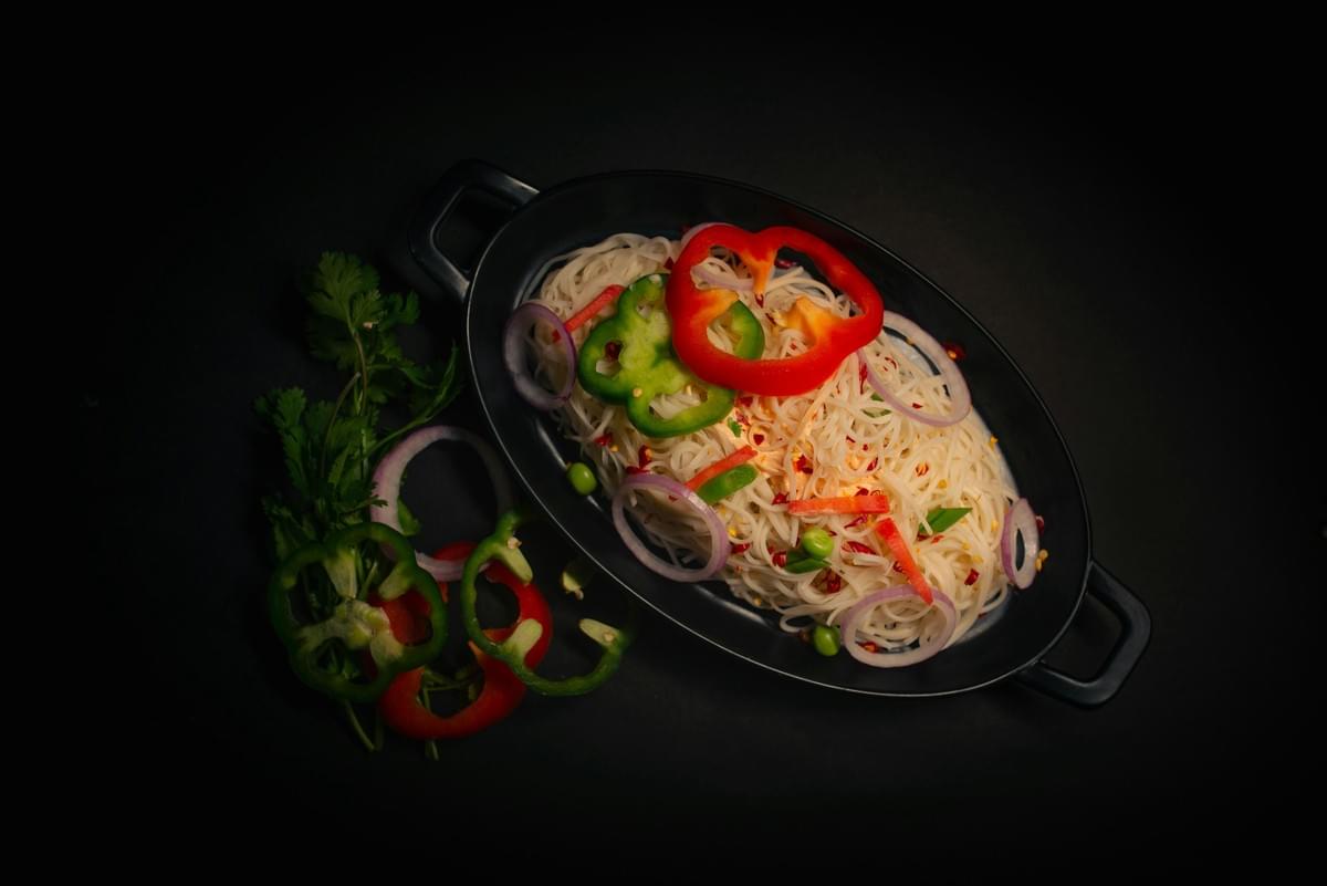 A black oval serving dish holds a bed of thin white noodles topped with vibrant slices of red and green bell peppers, red onion rings, green peas, and red chili flakes. The dish is set against a dark, moody background, flanked by fresh cilantro sprigs and additional sliced peppers on the side. A black oval serving dish holds a bed of thin white noodles topped with vibrant slices of red and green bell peppers, red onion rings, green peas, and red chili flakes. The dish is set against a dark, moody background, flanked by fresh cilantro sprigs and additional sliced peppers on the side.