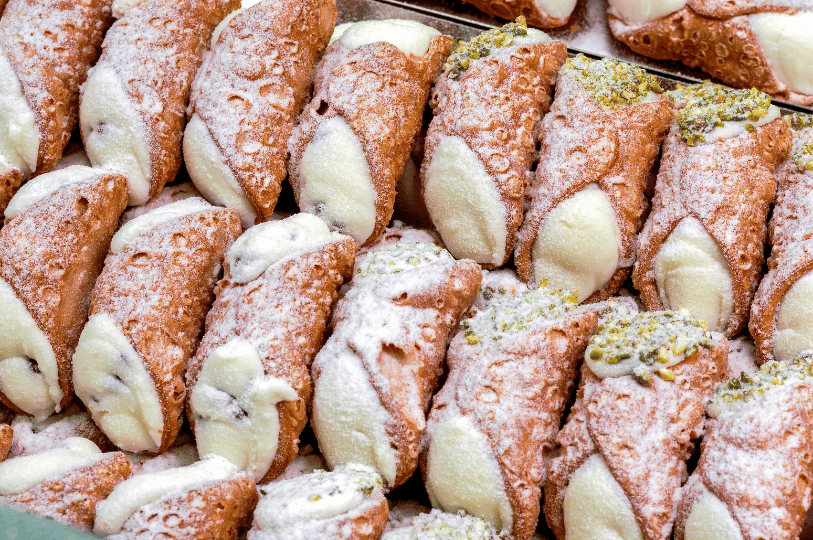 A high-angle, crowded display of numerous cannoli, some dusted heavily with powdered sugar and others garnished with crushed green pistachios on the cream ends. A high-angle, crowded display of numerous cannoli, some dusted heavily with powdered sugar and others garnished with crushed green pistachios on the cream ends.