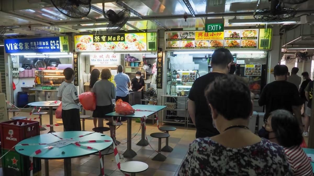 A bustling hawker center features people queueing at food stalls like "Ma Bo Lor Mee" and "Xing Wang Delights" for a meal. The dining area shows social distancing measures in place, with red and white hazard tape cordoning off sections of the circular tables. A bustling hawker center features people queueing at food stalls like "Ma Bo Lor Mee" and "Xing Wang Delights" for a meal. The dining area shows social distancing measures in place, with red and white hazard tape cordoning off sections of the circular tables.