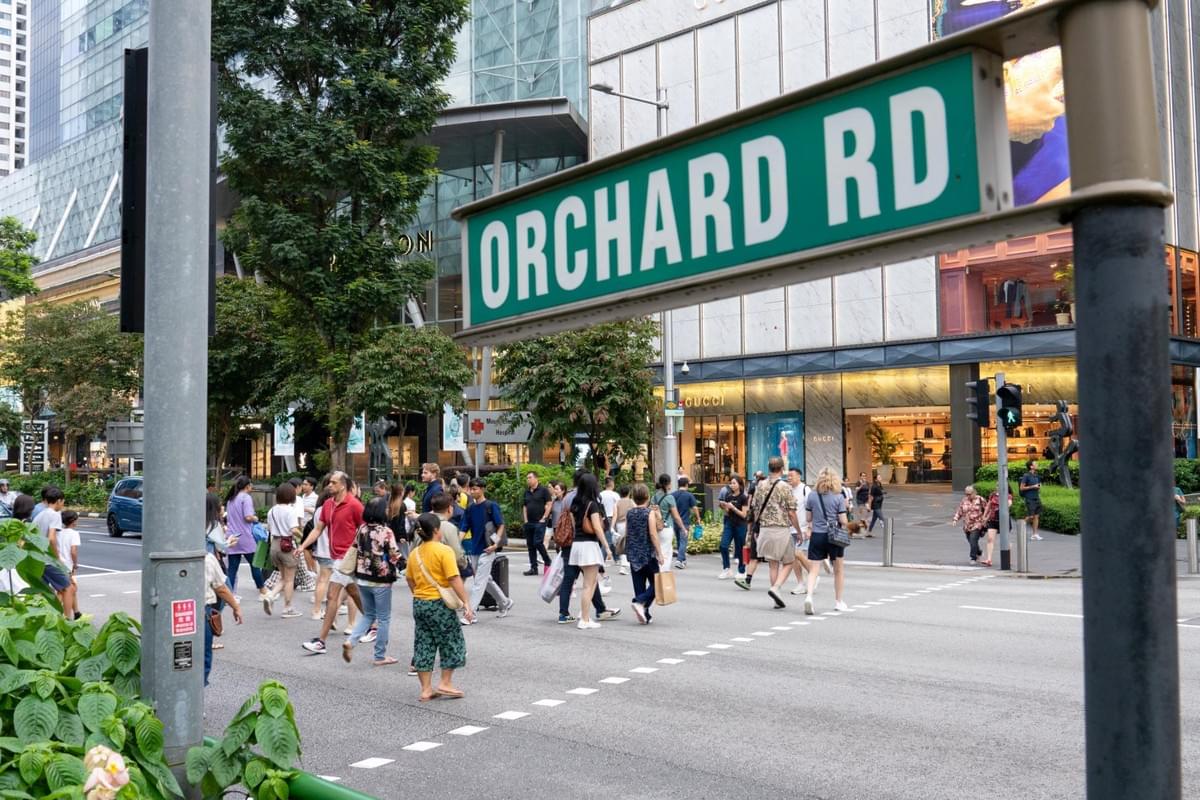 A green street sign reading "ORCHARD RD" stands prominently in the foreground at a busy intersection in Singapore. In the background, numerous pedestrians cross the street in front of high-end luxury retail stores like Gucci and Louis Vuitton. A green street sign reading "ORCHARD RD" stands prominently in the foreground at a busy intersection in Singapore. In the background, numerous pedestrians cross the street in front of high-end luxury retail stores like Gucci and Louis Vuitton.