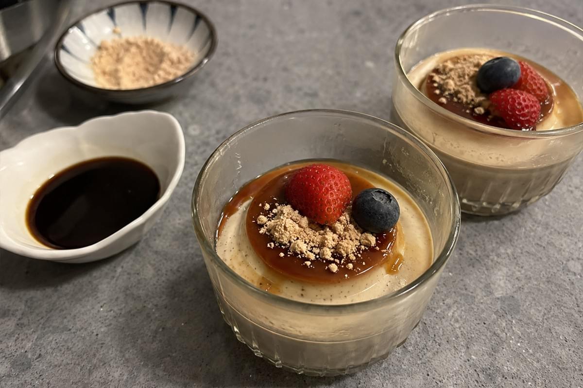 Two clear glass dishes filled with light brown hojicha panna cotta topped with a layer of glossy dark kuromitsu syrup, a dusting of kinako powder, a fresh strawberry piece, and a blueberry, arranged on a grey countertop next to small side bowls containing extra kinako powder and dark syrup. Two clear glass dishes filled with light brown hojicha panna cotta topped with a layer of glossy dark kuromitsu syrup, a dusting of kinako powder, a fresh strawberry piece, and a blueberry, arranged on a grey countertop next to small side bowls containing extra kinako powder and dark syrup.