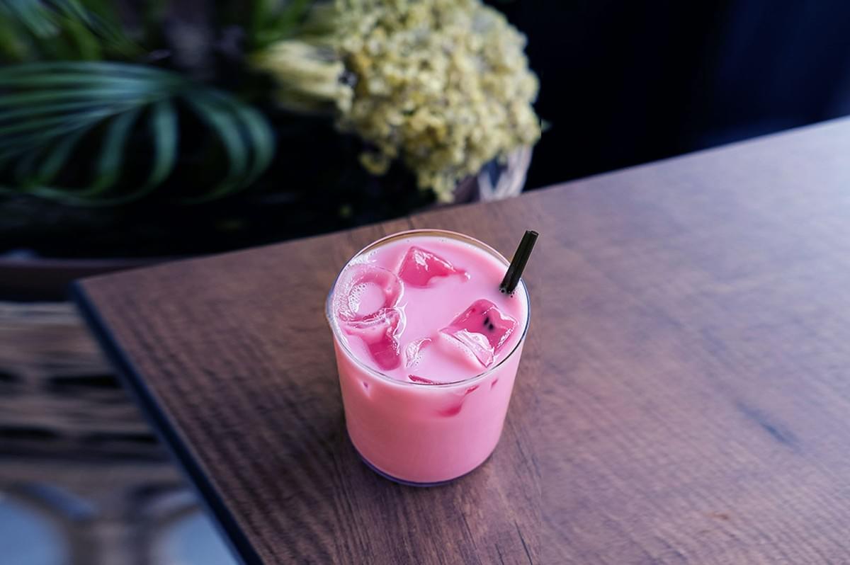 A glass of pink iced drink with a dark straw sits on a wooden table. Ice cubes float on top. Blurred greenery is visible in the background. A glass of pink iced drink with a dark straw sits on a wooden table. Ice cubes float on top. Blurred greenery is visible in the background.