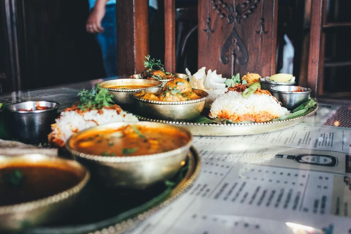 A traditional Indian Thali meal is served in brass bowls on large platters over a patterned table. The spread includes mounds of white rice, various curries, gravies, and small side condiments. A traditional Indian Thali meal is served in brass bowls on large platters over a patterned table. The spread includes mounds of white rice, various curries, gravies, and small side condiments.