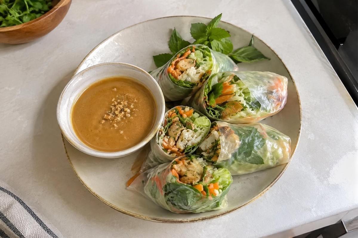 A high-angle view presents a large beige ceramic plate set on a white countertop, featuring fresh rice paper rolls cut in half to reveal fillings of cooked satay chicken strips, rice vermicelli, shredded carrots, cucumber, and fresh mint leaves. A small ceramic bowl containing smooth, peanut-free satay dip garnished with crushed seeds sits beside the rolls. A high-angle view presents a large beige ceramic plate set on a white countertop, featuring fresh rice paper rolls cut in half to reveal fillings of cooked satay chicken strips, rice vermicelli, shredded carrots, cucumber, and fresh mint leaves. A small ceramic bowl containing smooth, peanut-free satay dip garnished with crushed seeds sits beside the rolls.