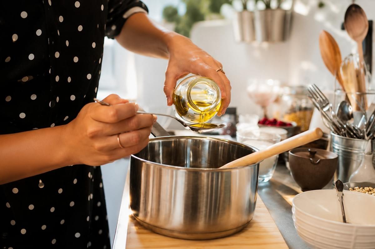 A person pouring golden olive oil from a small glass jar onto a metal spoon held over a stainless steel cooking pot. A person pouring golden olive oil from a small glass jar onto a metal spoon held over a stainless steel cooking pot.