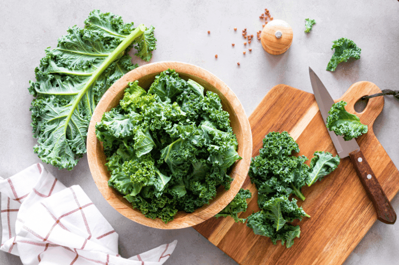A culinary flat-lay featuring a wooden bowl filled with chopped kale, a wooden cutting board with a knife, and scattered peppercorns on a gray stone surface. A culinary flat-lay featuring a wooden bowl filled with chopped kale, a wooden cutting board with a knife, and scattered peppercorns on a gray stone surface.