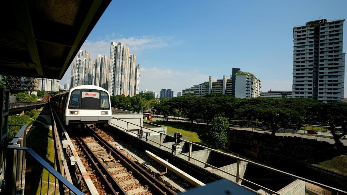A white SMRT train approaches an elevated station platform under a clear blue sky in Singapore. Tall residential apartment buildings and lush green trees line the tracks, creating a dense urban landscape. A white SMRT train approaches an elevated station platform under a clear blue sky in Singapore. Tall residential apartment buildings and lush green trees line the tracks, creating a dense urban landscape.