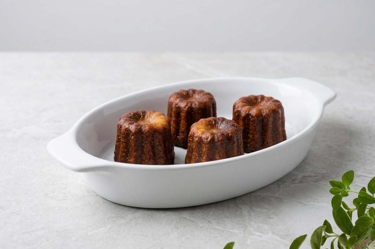 Four golden-brown canelés arranged in a white oval serving dish on a marble countertop Four golden-brown canelés arranged in a white oval serving dish on a marble countertop